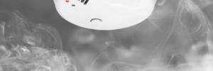 white smoke alarm mounted on a gray ceiling with smoke billowing around it